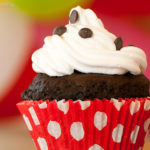 Close de um cupcake de chocolate com cobertura de chantilly e gotinhas de chocolate, em forminha vermelha com poás brancos.
