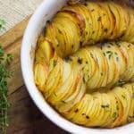 Prato de batata laminada crocante assada, disposta em camadas finas e douradas, temperada com manteiga e ervas frescas, servida em uma assadeira branca sobre uma tábua de madeira.