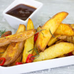 Travessa branca com batatas rústicas douradas e crocantes, temperadas com azeite, alecrim e pimenta.