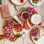 Tábua de queijos decorada com uvas, maçãs, alecrim, embutidos e uma pequena tigela de geleia vermelha, compondo um cenário acolhedor e festivo.
