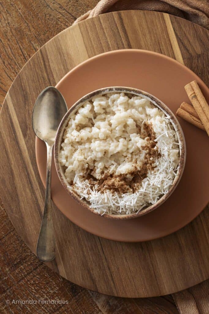 Vista de cima de tigela de arroz doce com coco e canela ao lado de pau de canela e pano de fundo marrom.