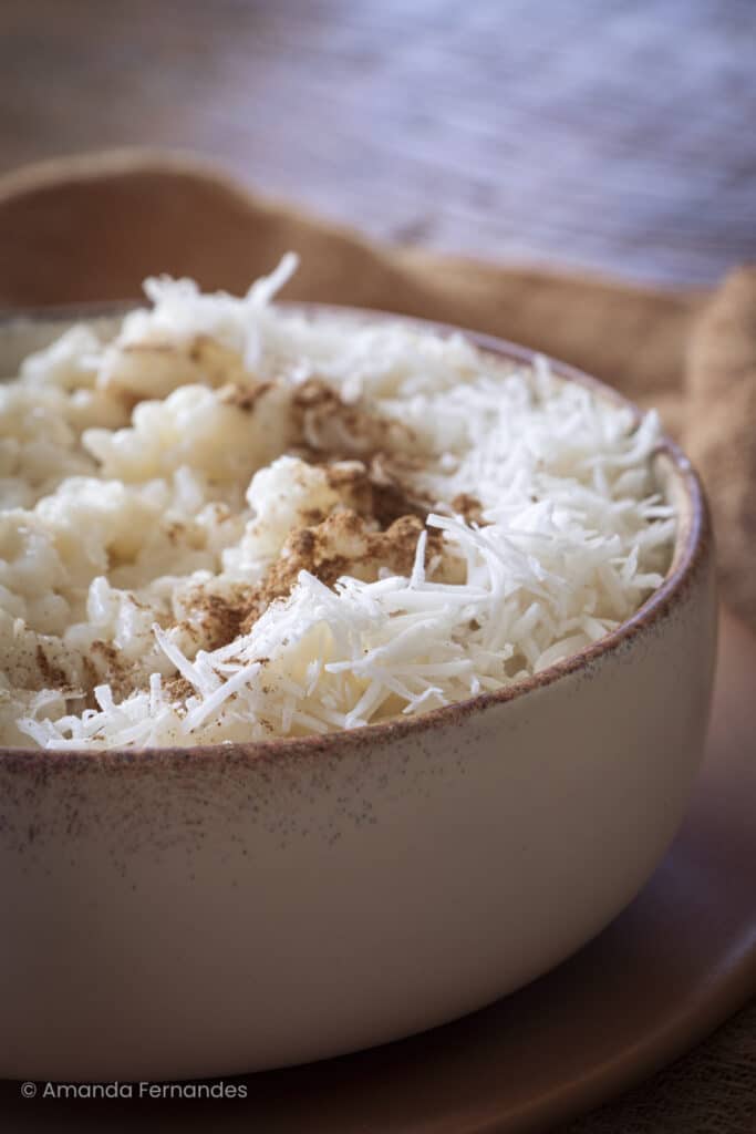Porção de arroz doce cremoso em tigela clara, finalizado com coco ralado e toque de canela, sobre pano de linho.