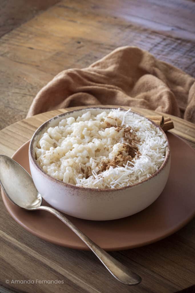 Tigela de arroz doce cremoso com coco ralado e canela, sobre prato rústico de cerâmica em fundo de madeira.